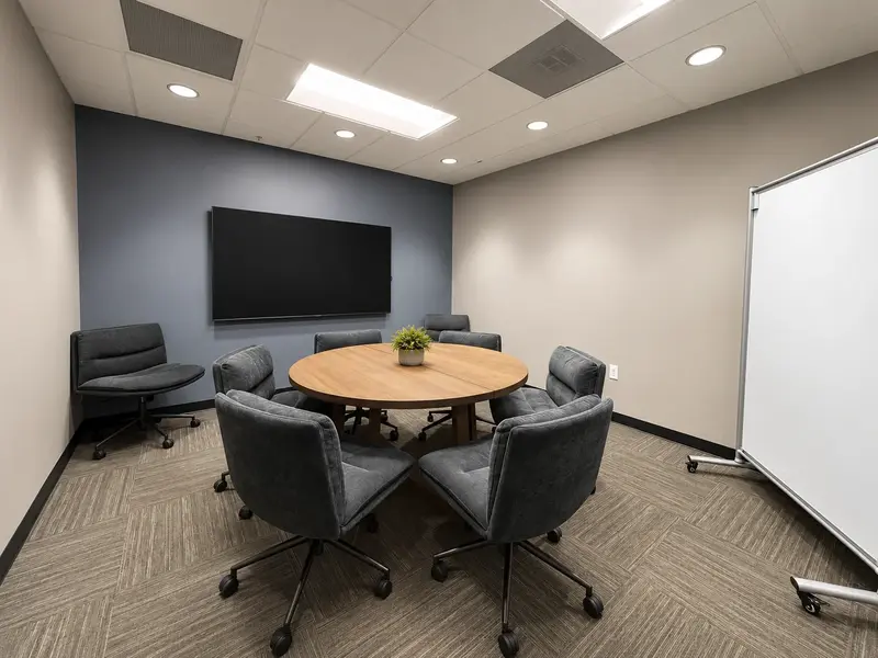 The Conference Lounge meeting room featuring a round wooden table, plush chairs, display screen, and whiteboard.