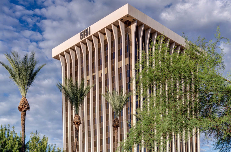 Exterior view of the 5151 E Broadway Blvd office building in Tucson, Arizona, framed by palm trees under a cloudy sky.