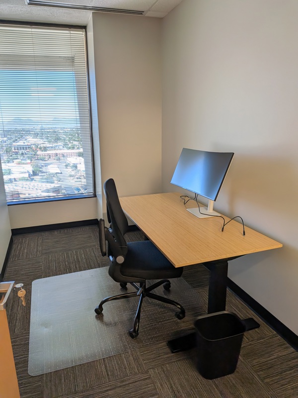 Private office Suite 155 in Tucson with a bamboo sit-stand desk, curved monitor, and city views through large windows.