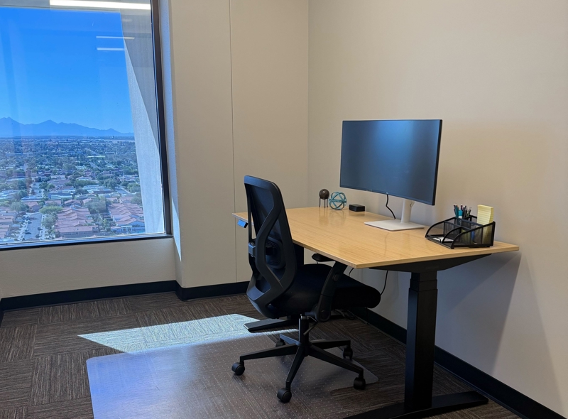 Modern private office with an adjustable sit-stand desk and ergonomic chair featuring a large window with city views.