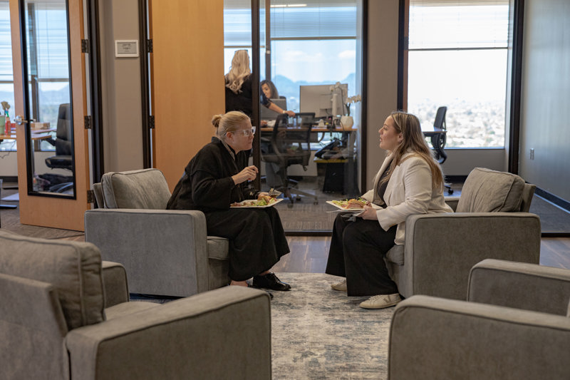 Two women having a conversation over lunch in a comfortable office lounge area with city views in the background.