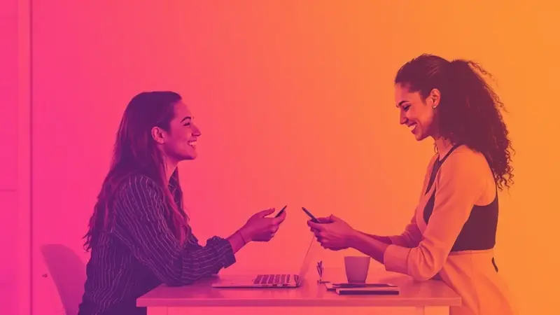 Two women smiling and using smartphones while coworking at a desk with a laptop under a pink and orange gradient.