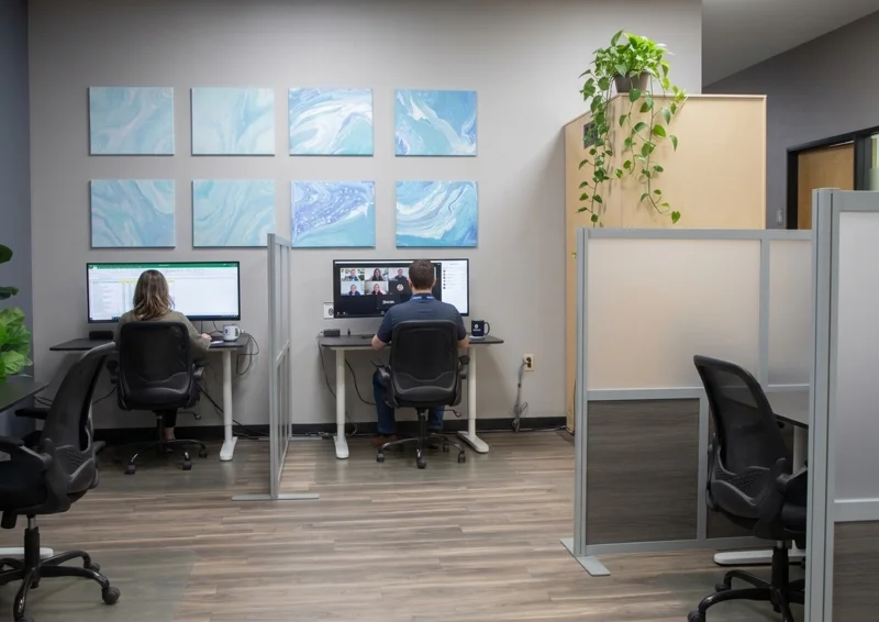 Modern coworking space featuring two hot desk workstations with monitors, ergonomic chairs, and privacy screens.