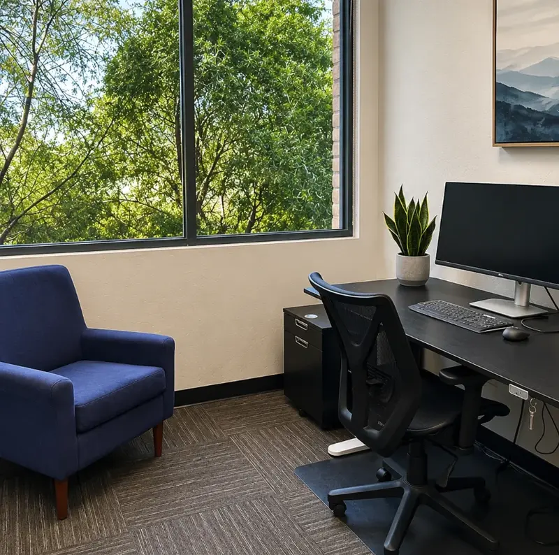 Private office Suite 122 at Spoke Coworking with sit-stand desk, curved monitor, armchair, and lush green window views.